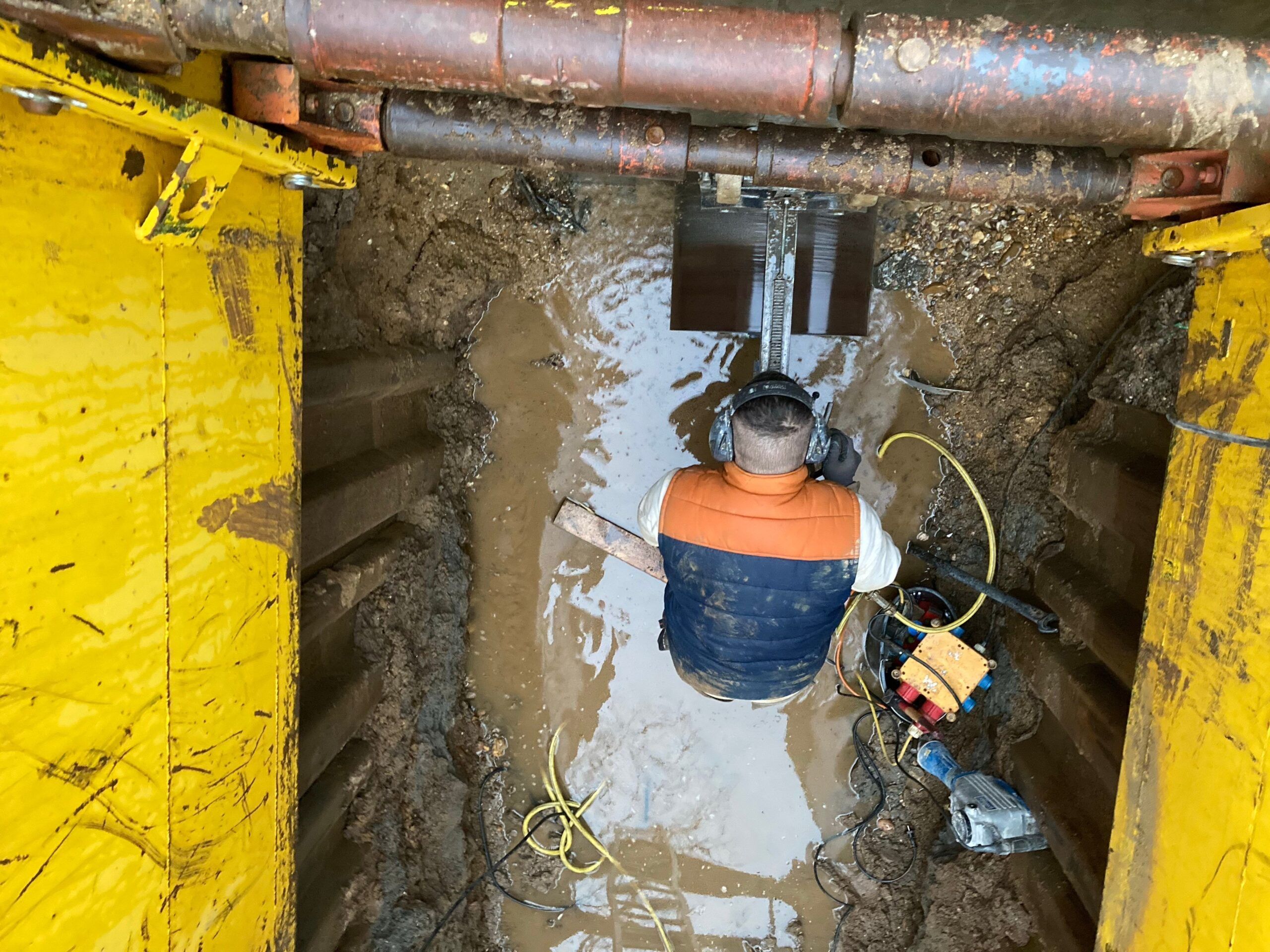 Blick von oben in einen Graben mit gelbem Verbau, ein Mitarbeiter mit Gehörschutz arbeitet an der Grabenwand